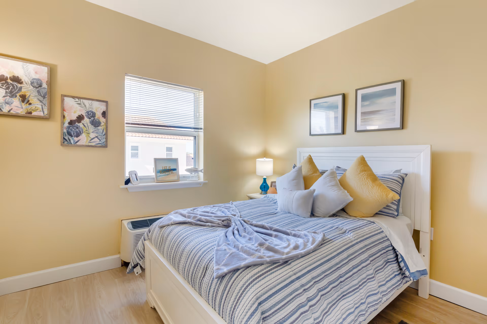Bright bedroom with a striped bed topped with multiple pillows, framed artwork, a window and a bedside lamp.