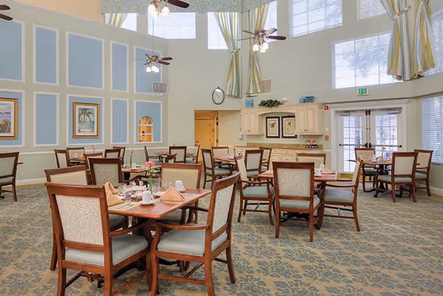 A bright dining room with multiple wooden tables and chairs arranged neatly. Each table is set with napkins, cups, and small flower vases. The room features high ceilings with large windows covered by striped curtains, ceiling fans with lights, and light blue walls with white trim. There is a small kitchenette area with cabinets and framed artwork on the walls.