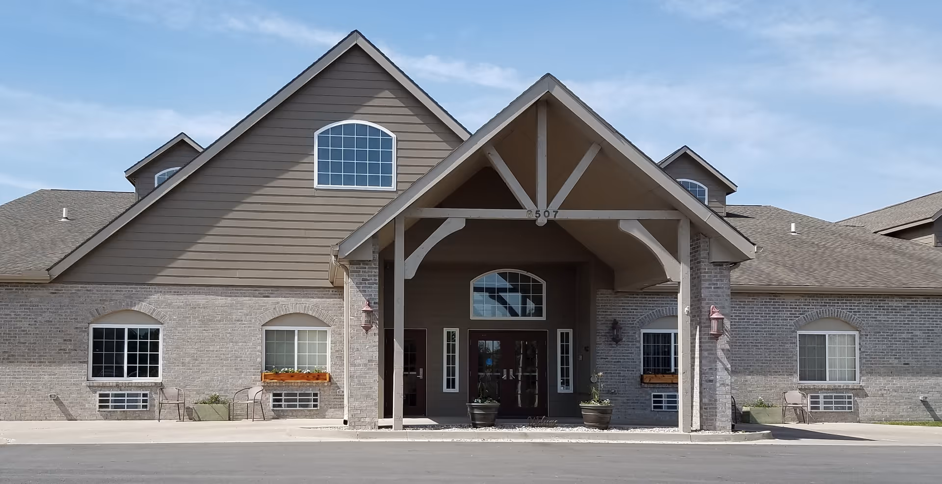Front exterior view of a single-story assisted living facility building with a peaked roof and large covered entrance. The building has light gray brick walls, multiple windows with white frames, flower boxes, and outdoor chairs near the entrance. The sky is clear and blue.