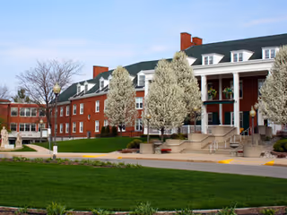 Exterior view of a large brick building with white columns and multiple windows, surrounded by blooming trees and green grass under a clear sky.