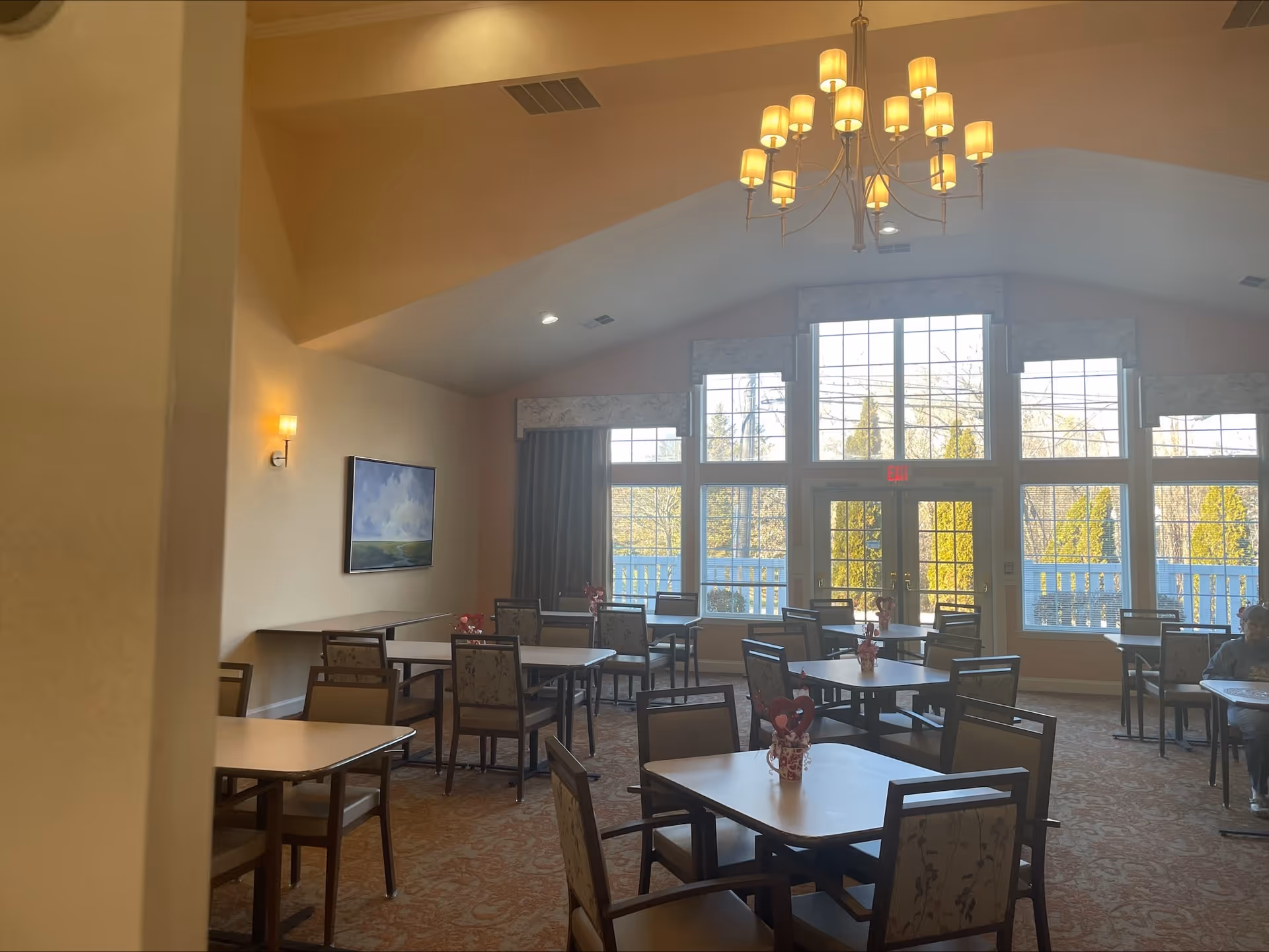 A dining room with multiple square tables and chairs arranged neatly. Each table has a small decorative centerpiece. Large windows with curtains allow natural light to fill the room. A chandelier with multiple lights hangs from the ceiling. There is a painting on the left wall and a person sitting at a table on the right side of the room.