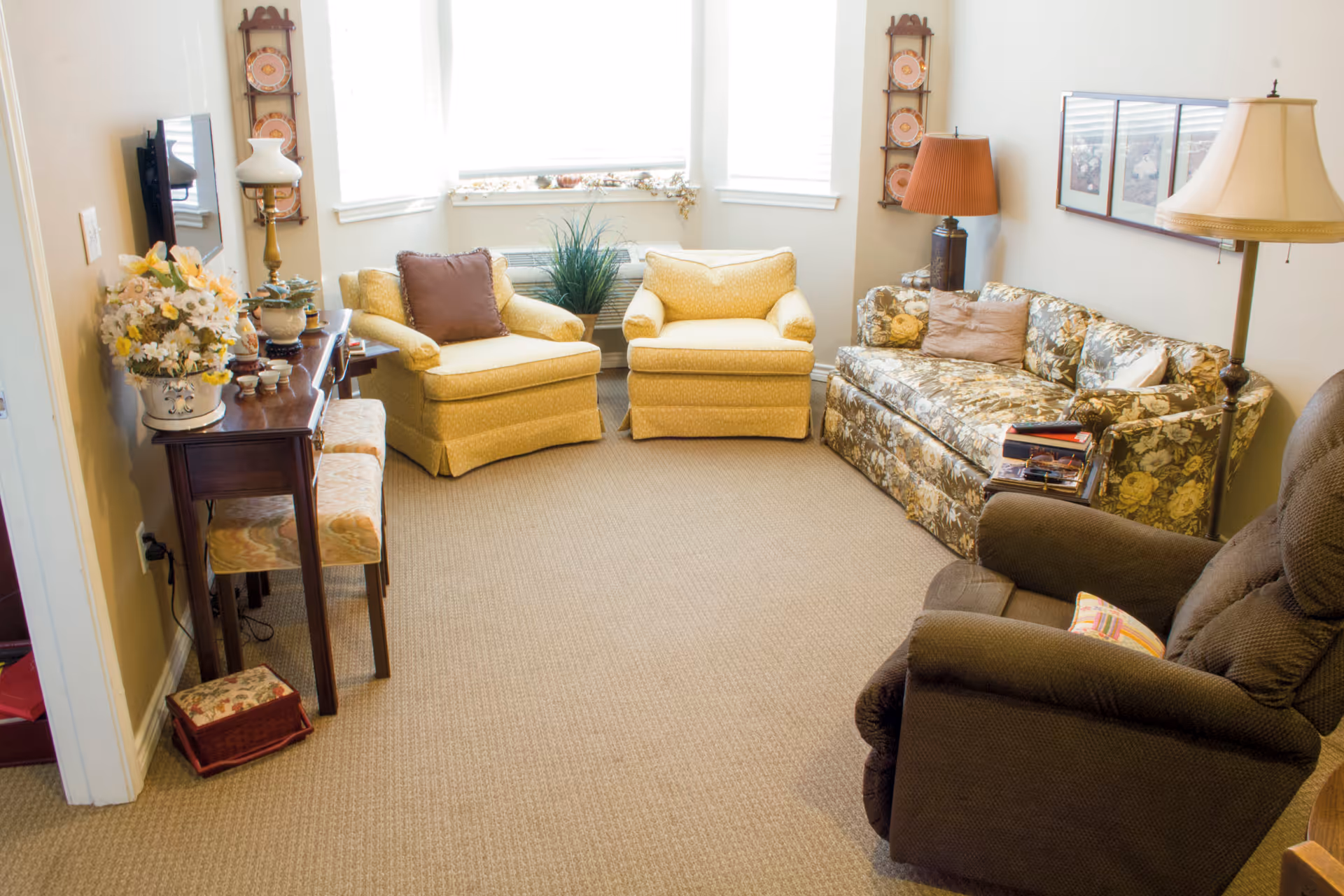A cozy living room with a floral-patterned sofa, two yellow armchairs, a brown recliner, and a wooden console table with decorative items and flowers. The room is well-lit by natural light coming through three windows with white blinds.