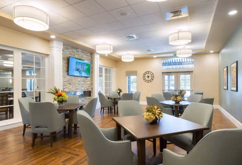 A well-lit common area with several square wooden tables surrounded by cushioned chairs. Each table has a floral centerpiece. The room features a stone accent wall with a mounted flat-screen TV above a modern fireplace. There are large windows and glass doors letting in natural light, a wall clock, and framed artwork on the walls. The ceiling has multiple round light fixtures.