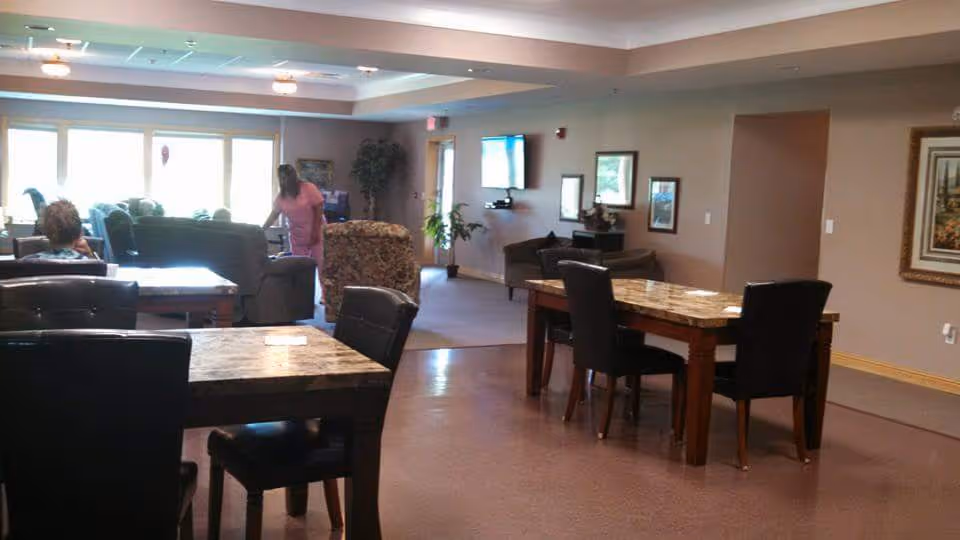 Interior view of a senior living facility common area with multiple tables and chairs, a few armchairs, a wall-mounted TV, framed pictures on the walls, and a person standing near the seating area. The room is well-lit with natural light coming from large windows.
