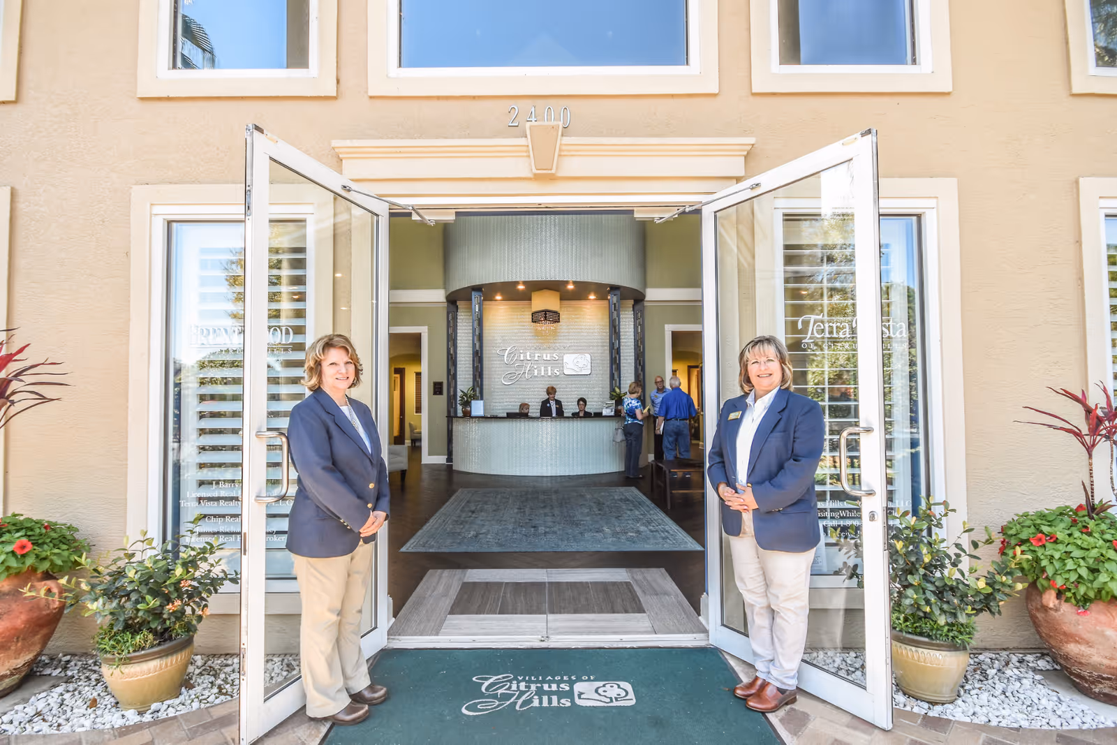 Two women in navy blazers and beige pants stand at the open glass doors of the entrance to The Villages of Citrus Hills facility. The entrance features potted plants on either side and a green welcome mat with the facility's name. Inside, a reception desk with staff and visitors is visible.