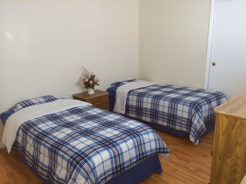 A simple bedroom with two twin beds covered in blue and white plaid bedding. Between the beds is a small wooden nightstand with a vase of pink flowers. The room has light-colored walls, a wooden floor, a wooden dresser on the right side, and a closed white door in the background.