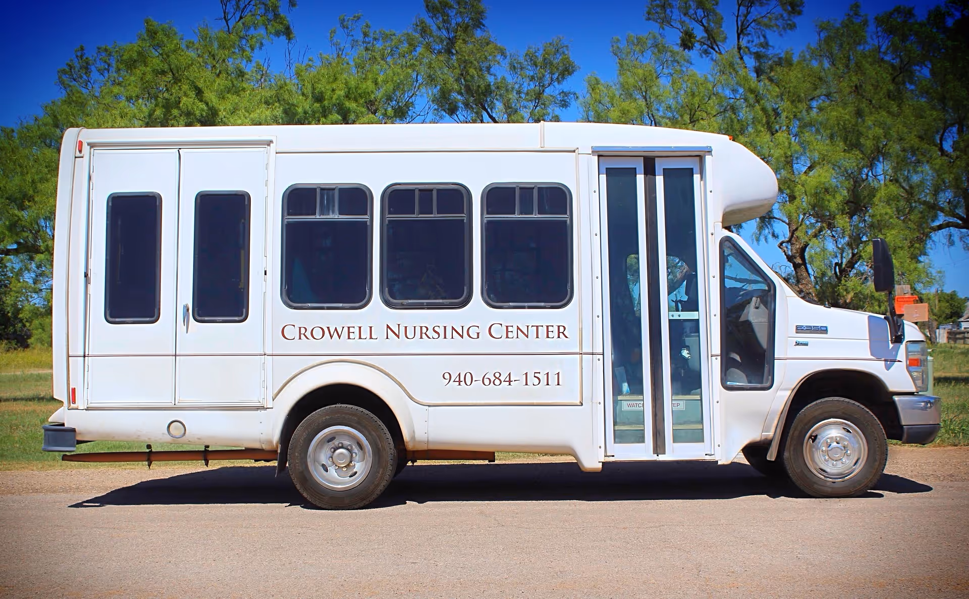 White shuttle bus parked on a road with trees and blue sky in the background. The side of the bus displays the text 'Crowell Nursing Center' and a phone number '940-684-1511'.