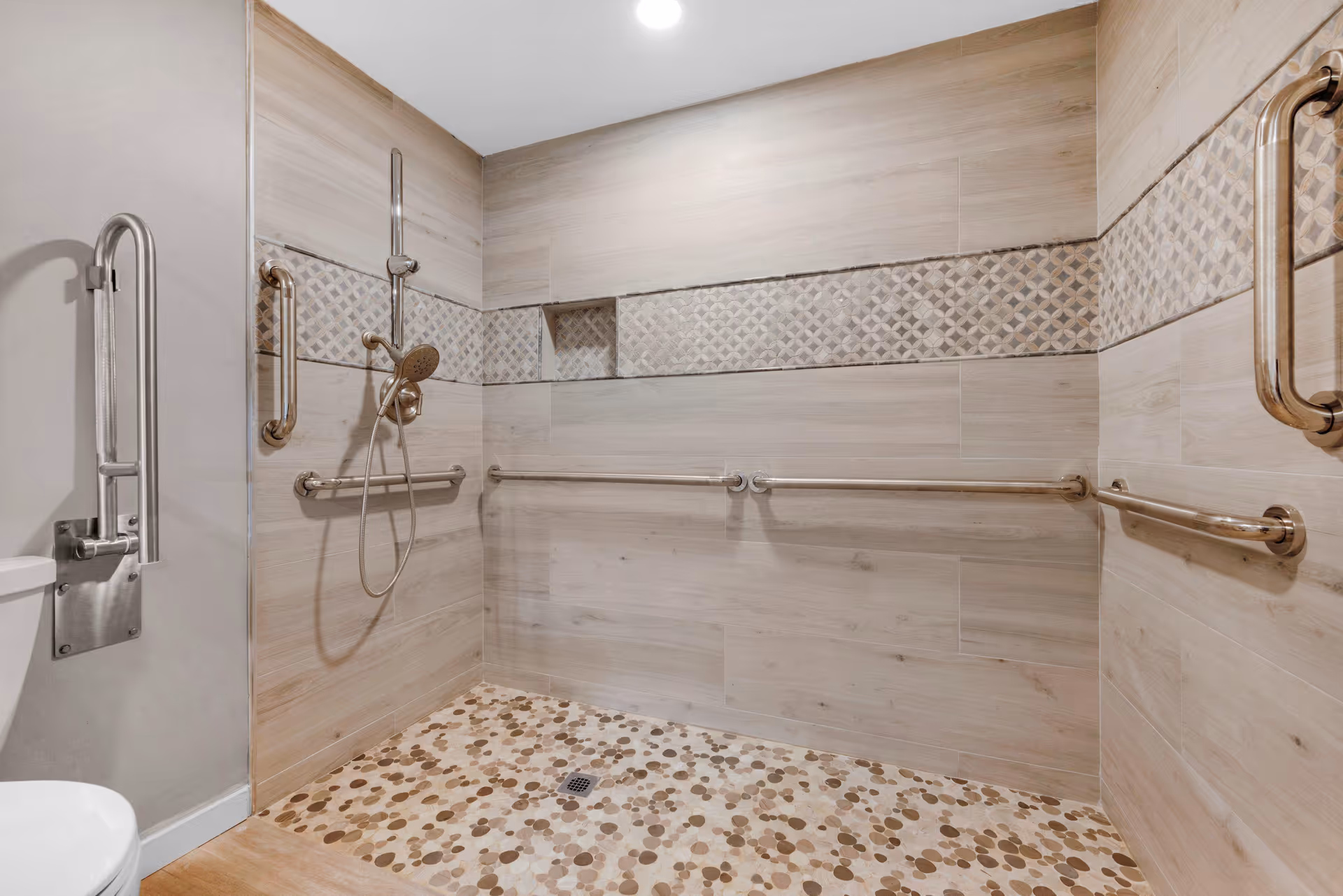 Accessible shower area with multiple stainless steel grab bars mounted on light wood-patterned tiled walls. The shower floor has a pebble-like tile design in various shades of brown. A handheld showerhead is attached to the wall, and a small recessed shelf is visible in the tiled wall. Part of a toilet with a support bar is visible on the left side.