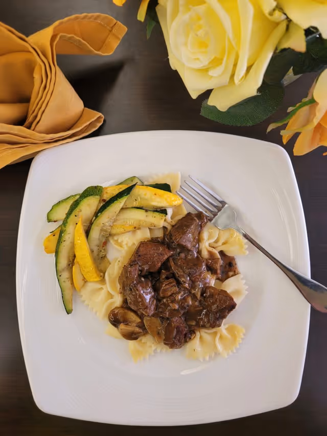 A white square plate with beef stew served over bowtie pasta, accompanied by sautéed zucchini and yellow squash. A fork rests on the plate, and a folded mustard-colored napkin and yellow roses are visible on the dark table surface.