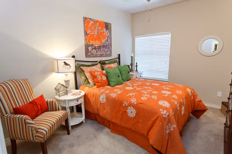 Sunny bedroom with an orange floral bedspread, green pillows, a striped armchair, nightstand with a lamp, and wall art.