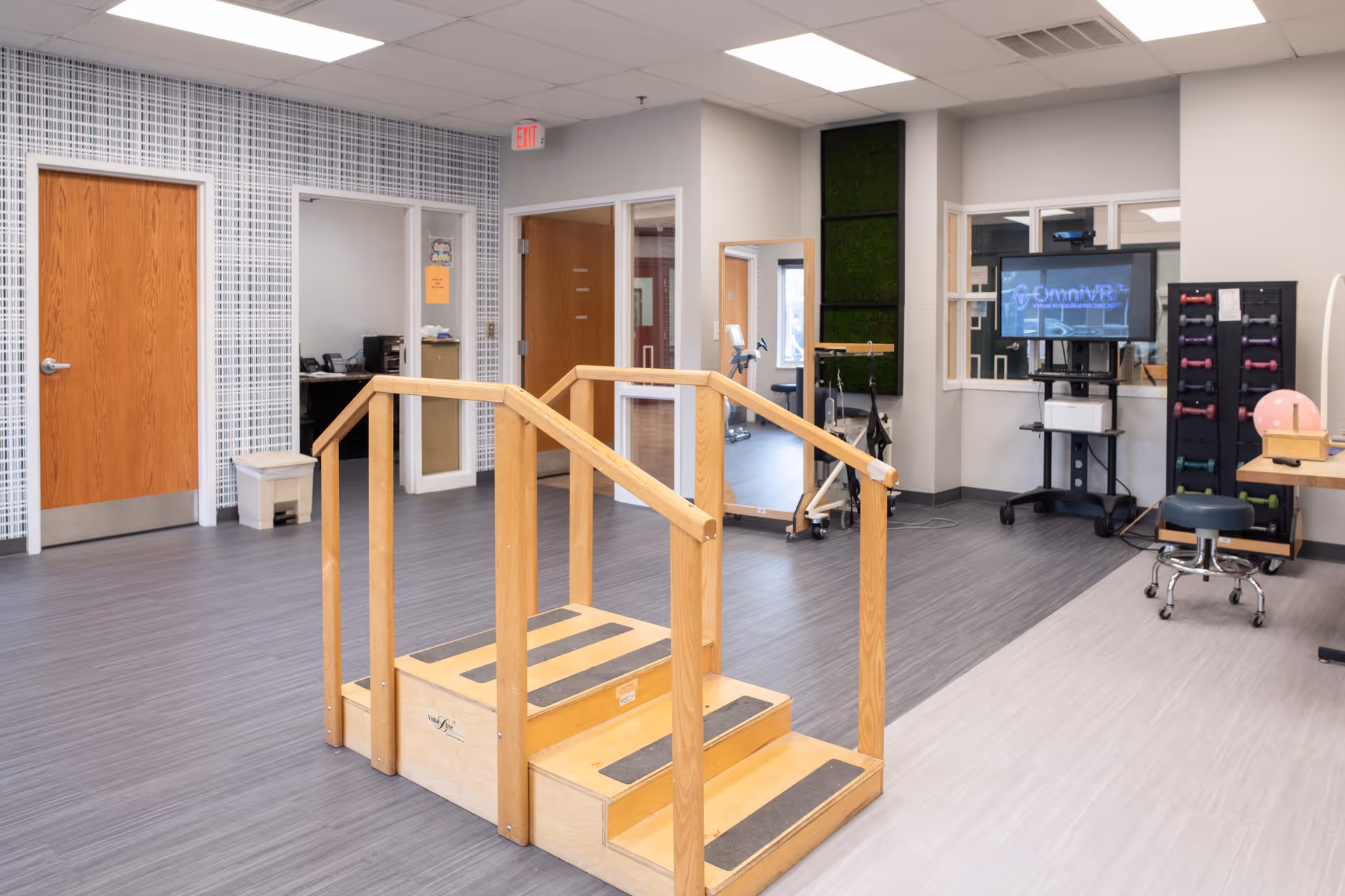 A rehabilitation or physical therapy room with a wooden practice staircase with handrails in the center. The room has gray flooring, light gray walls, and wooden doors. There is exercise equipment including a rack of colorful dumbbells, a pink exercise ball, a stool, and a TV screen mounted on a stand displaying a virtual reality interface. A large mirror and a window looking into another room are also visible.