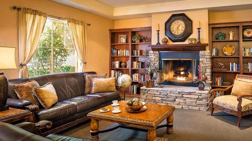 Cozy living room with a dark leather sofa and an upholstered armchair around a wooden coffee table. The room features a stone fireplace with a fire burning, a large clock above the mantel, and built-in wooden bookshelves filled with books and decorative items. Light curtains frame a window letting in natural light.