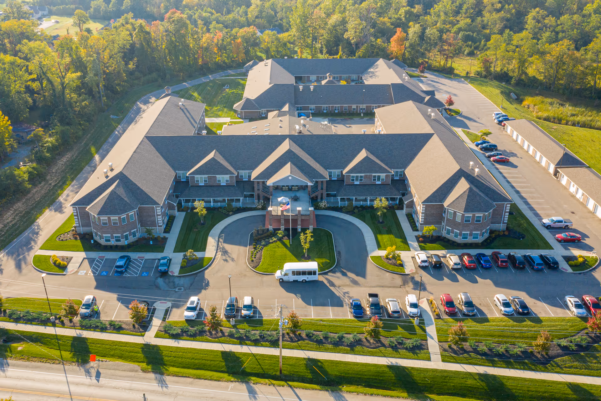 Aerial view of Magnolia Springs Loveland, a large senior living facility surrounded by greenery. The building has multiple wings with a central entrance featuring a covered drop-off area. There are several cars parked in the parking lots around the building, and a white shuttle van is parked near the entrance. The facility is set in a scenic area with trees and open land in the background.