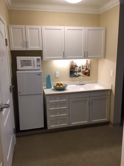 Small kitchenette with white cabinets, a countertop sink, compact refrigerator and microwave, and a bowl of fruit.