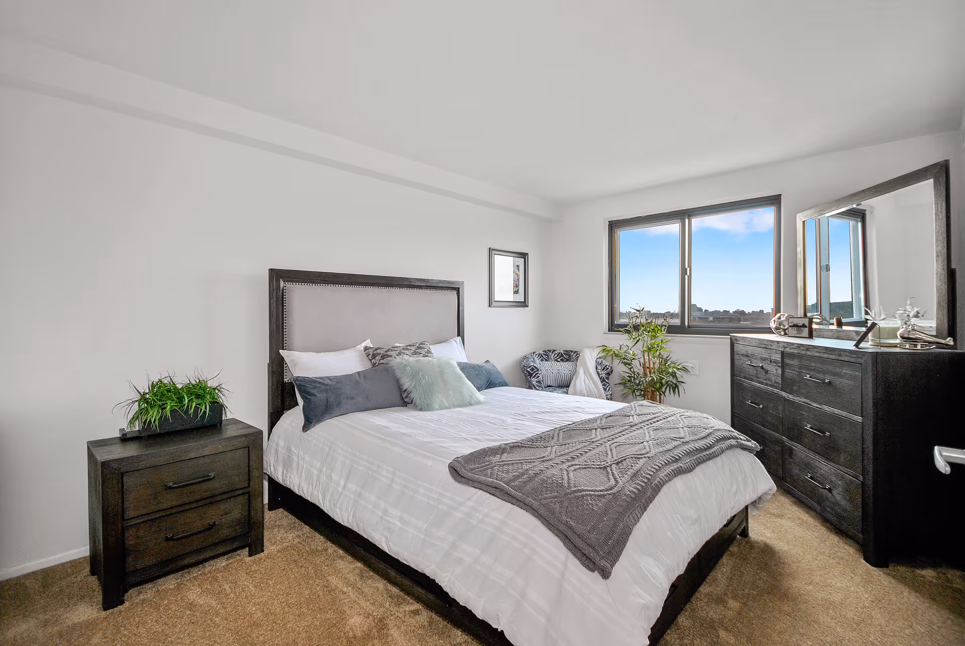 A bright bedroom featuring a neatly made bed, dark wood nightstand and dresser, a window, and potted plants.