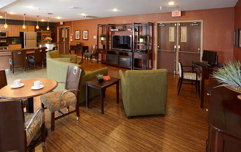 A spacious and well-lit common area in Canterbury Nursing and Rehabilitation Center featuring a living room and kitchen. The living room has green upholstered chairs, a wooden coffee table, a TV on a wooden entertainment center, and framed artwork on the walls. The kitchen area includes a refrigerator, oven, microwave, and a counter with bar stools. There are also small round tables with chairs and a computer desk with a chair near double exit doors.