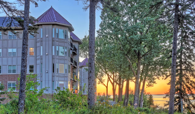 Exterior view of a multi-story senior living facility building with a purple roof, surrounded by tall trees and greenery, with a sunset over a body of water in the background.