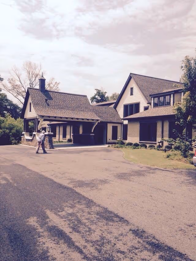 Exterior view of a large residential-style building with a covered entrance and multiple windows, surrounded by trees and greenery. A person is walking on the driveway leading to the entrance under a partly cloudy sky.