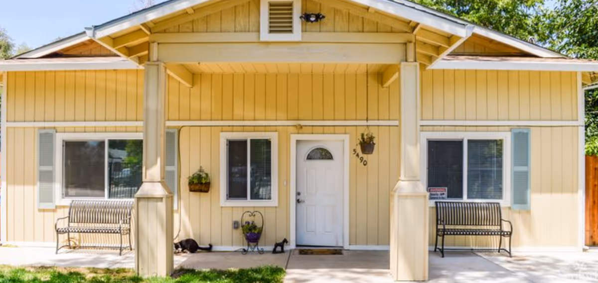 Front exterior view of a single-story yellow building with a white door and two windows, each with light blue shutters. There are two black metal benches on either side of the door, a hanging plant to the left of the door, and a small potted plant on the ground. The building has a covered porch supported by two columns.