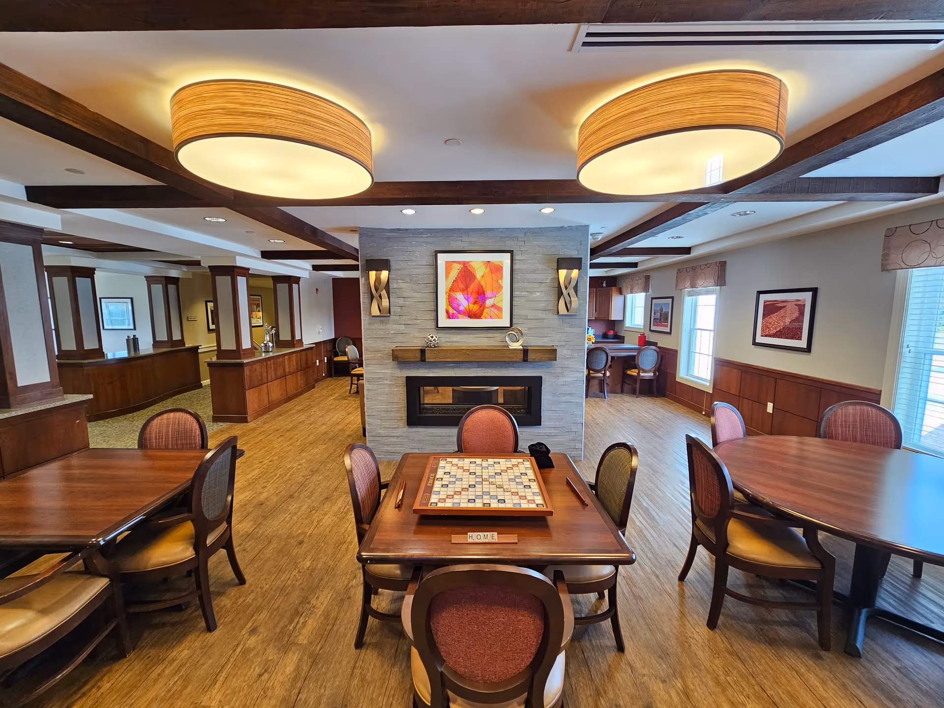 A spacious and well-lit common area in a senior living facility featuring wooden tables and chairs arranged for social activities. In the center, there is a table with a Scrabble board game and letter tiles spelling 'HOME'. The room has a modern fireplace with decorative wall sconces and a colorful framed artwork above it. Large round ceiling lights and wooden ceiling beams add warmth to the space. Windows with blinds allow natural light to enter, and framed pictures decorate the walls.