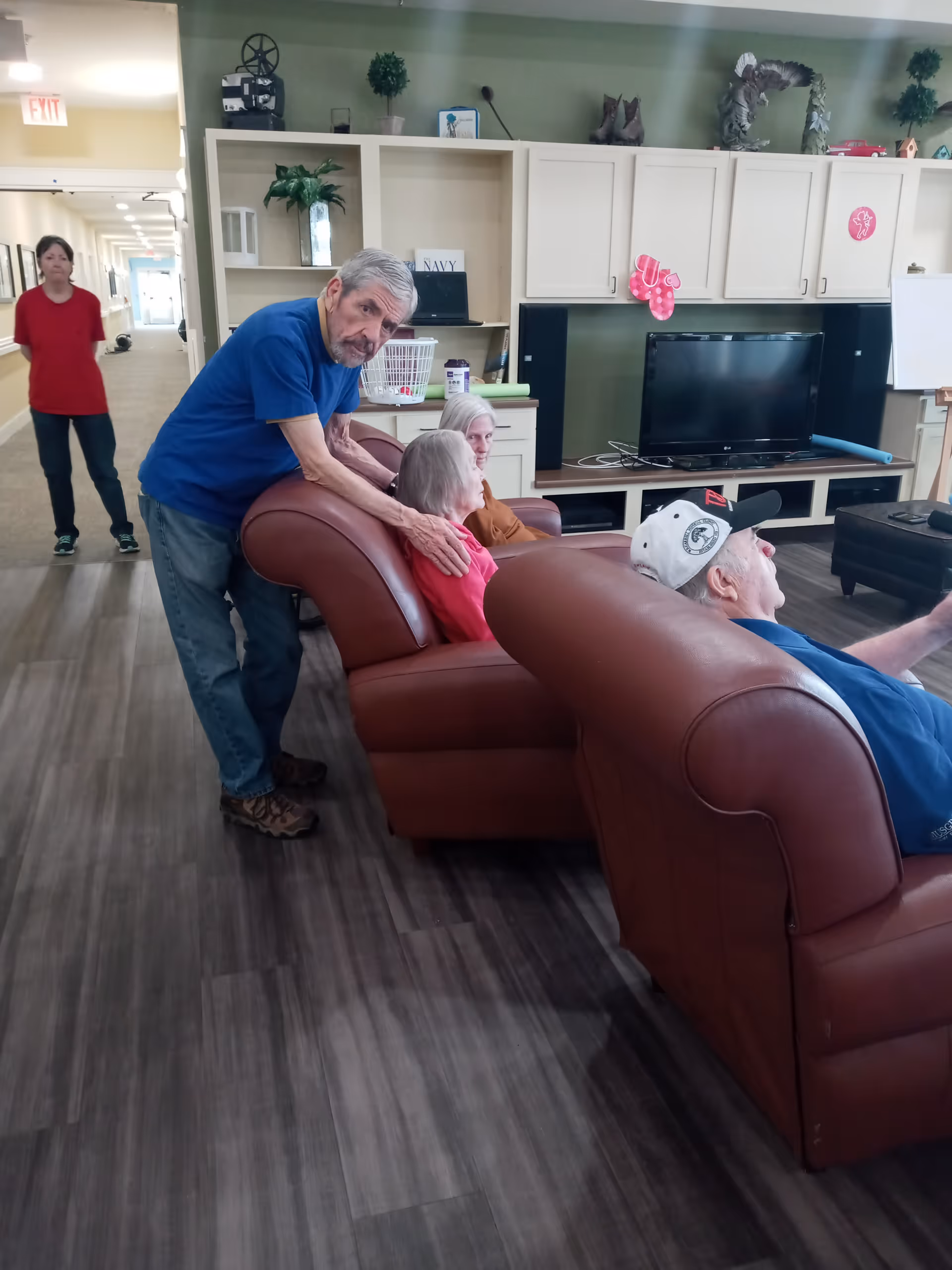 A group of elderly people sitting and standing around brown leather couches in a common living area with a TV, shelves, and decorations. A man in a blue shirt is standing and leaning on the couch, while two women and another man are seated. A woman in a red shirt stands in the hallway in the background.