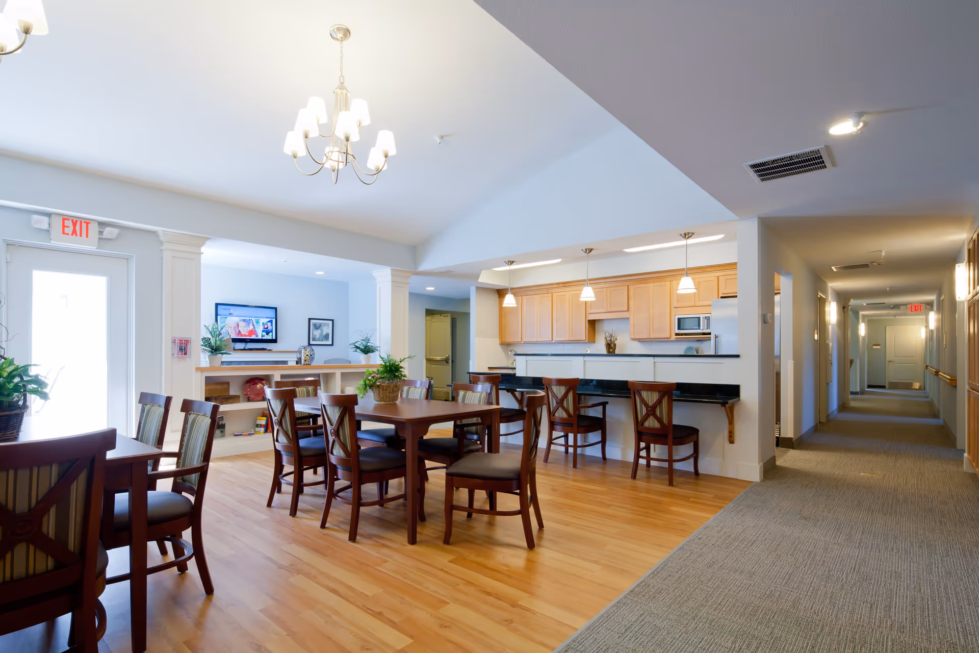 A bright and spacious common area in a senior living facility featuring a dining table with chairs, a kitchen area with wooden cabinets and a breakfast bar with stools, and a hallway leading to other rooms. The room has hardwood flooring, pendant lights above the kitchen counter, and a TV mounted on the wall with plants and decorative items on shelves below it.