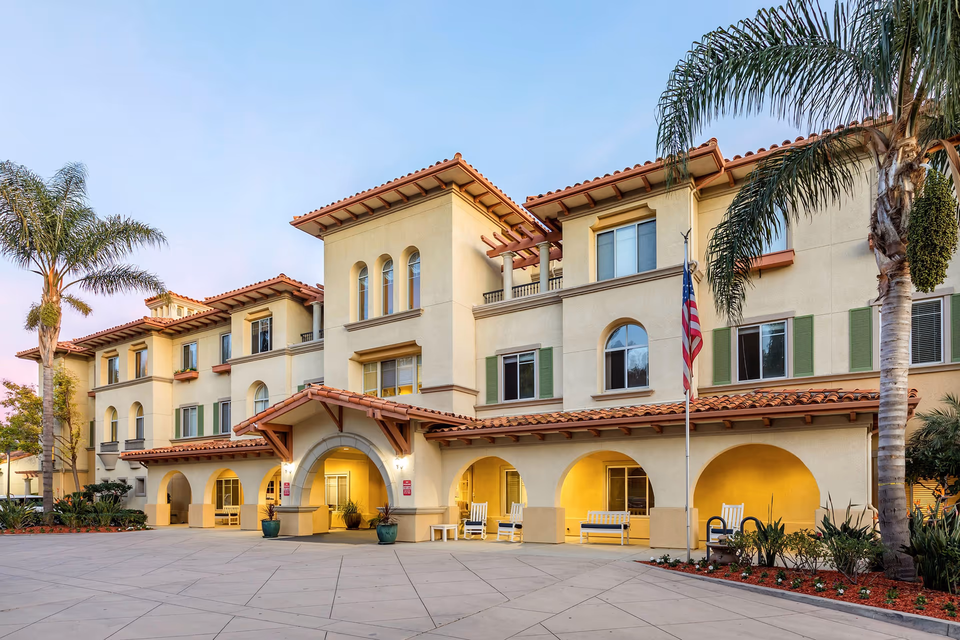 Exterior view of a three-story senior living facility building with a Mediterranean architectural style, featuring beige walls, red tile roofs, arched entryways, and several windows. There are palm trees and landscaped plants around the building, an American flag on a flagpole, and outdoor seating with benches and rocking chairs under the arches.