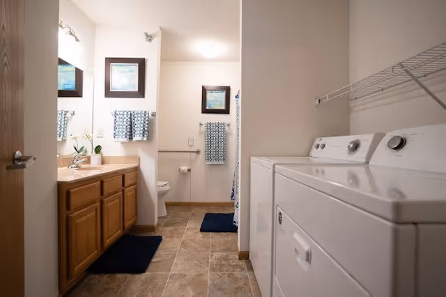 A combined bathroom and laundry area featuring a wooden vanity with a sink and mirror on the left, a toilet and shower with blue patterned towels and a framed picture on the wall in the center, and a white washing machine and dryer on the right with a wire shelf above them. The floor is tiled and there are dark blue rugs placed in front of the vanity and shower.