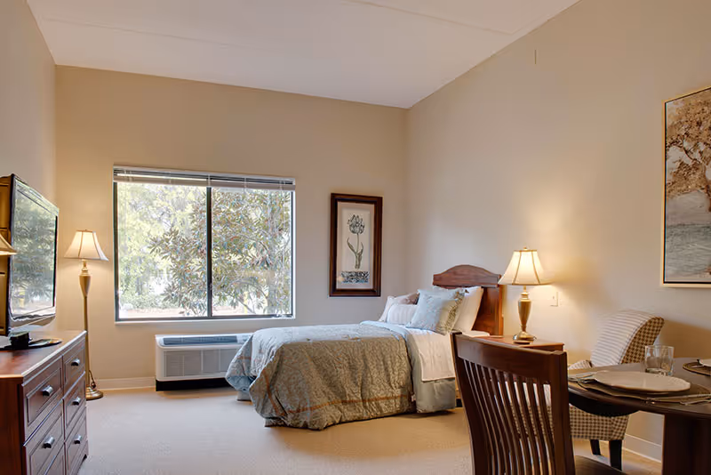 A cozy senior living bedroom with a neatly made bed, a wooden dresser with a TV on top, a floor lamp, a window showing greenery outside, a small dining table with two chairs, and framed artwork on the walls.