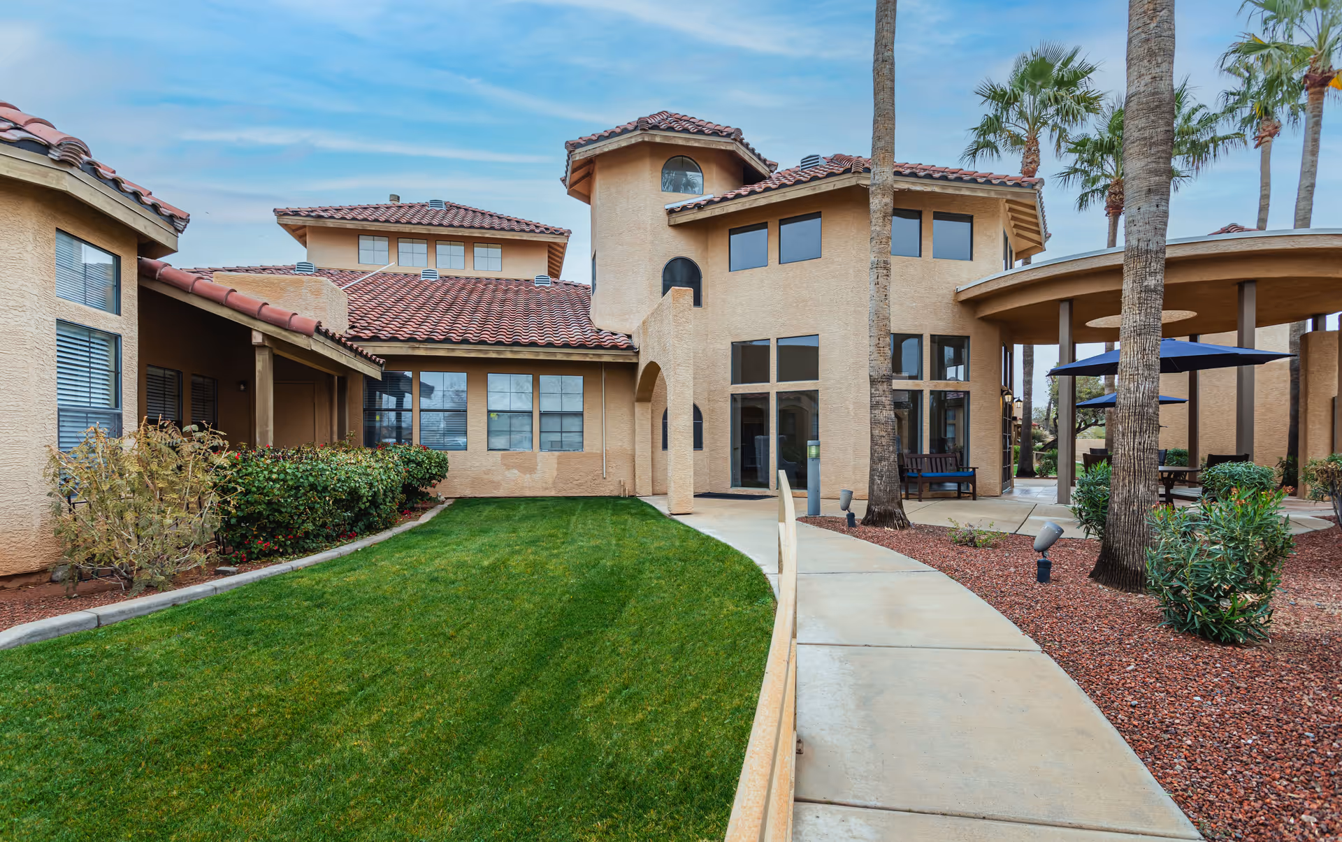 Exterior view of a senior living facility building with beige stucco walls and red tile roofs. The scene includes a curved concrete walkway, green lawn, palm trees, and a covered outdoor seating area with benches and umbrellas.