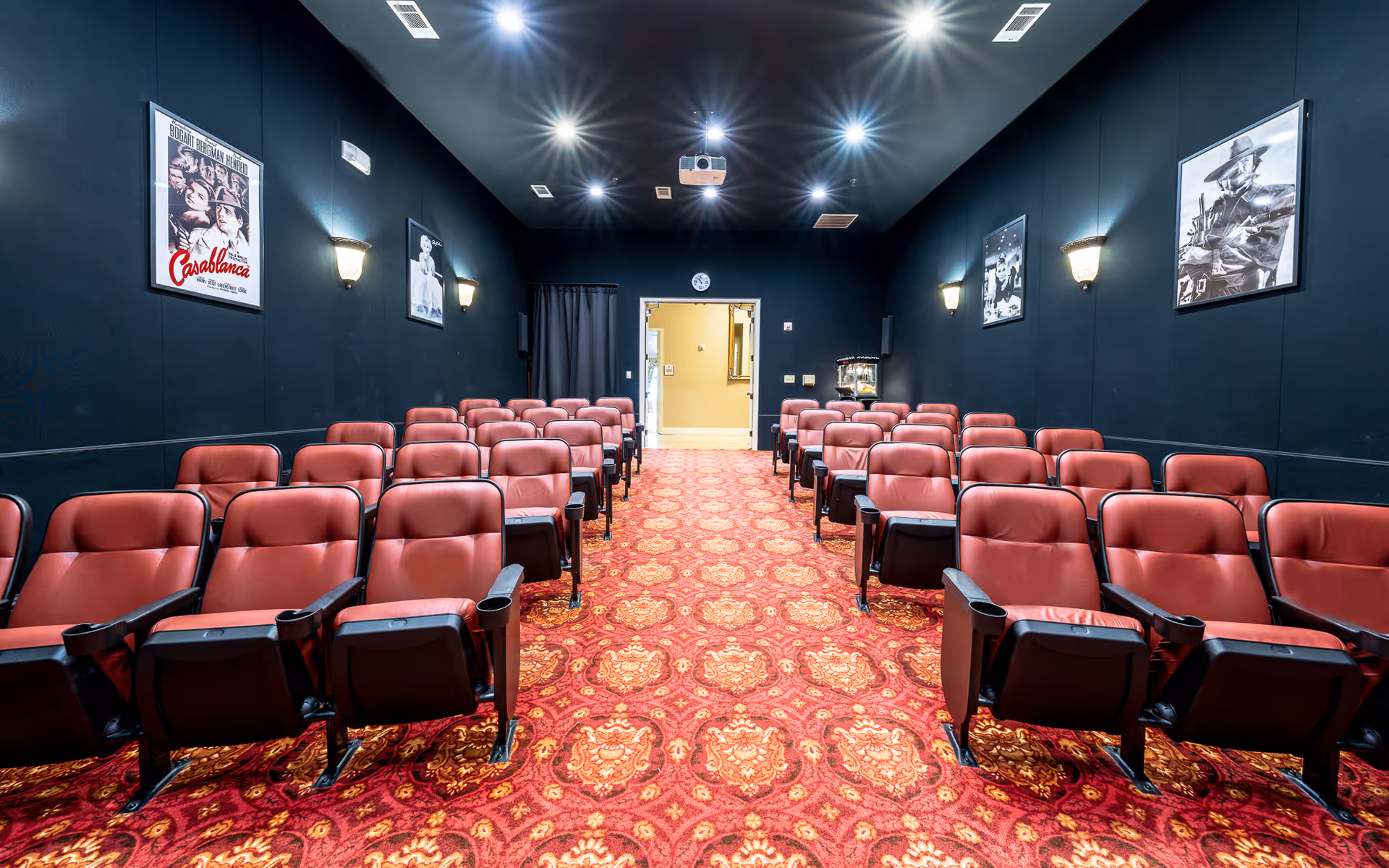 Interior view of a small movie theater room with rows of red cushioned seats, a red patterned carpet, dark walls adorned with classic movie posters, and ceiling lights illuminating the space. A projector is mounted on the ceiling at the back of the room.