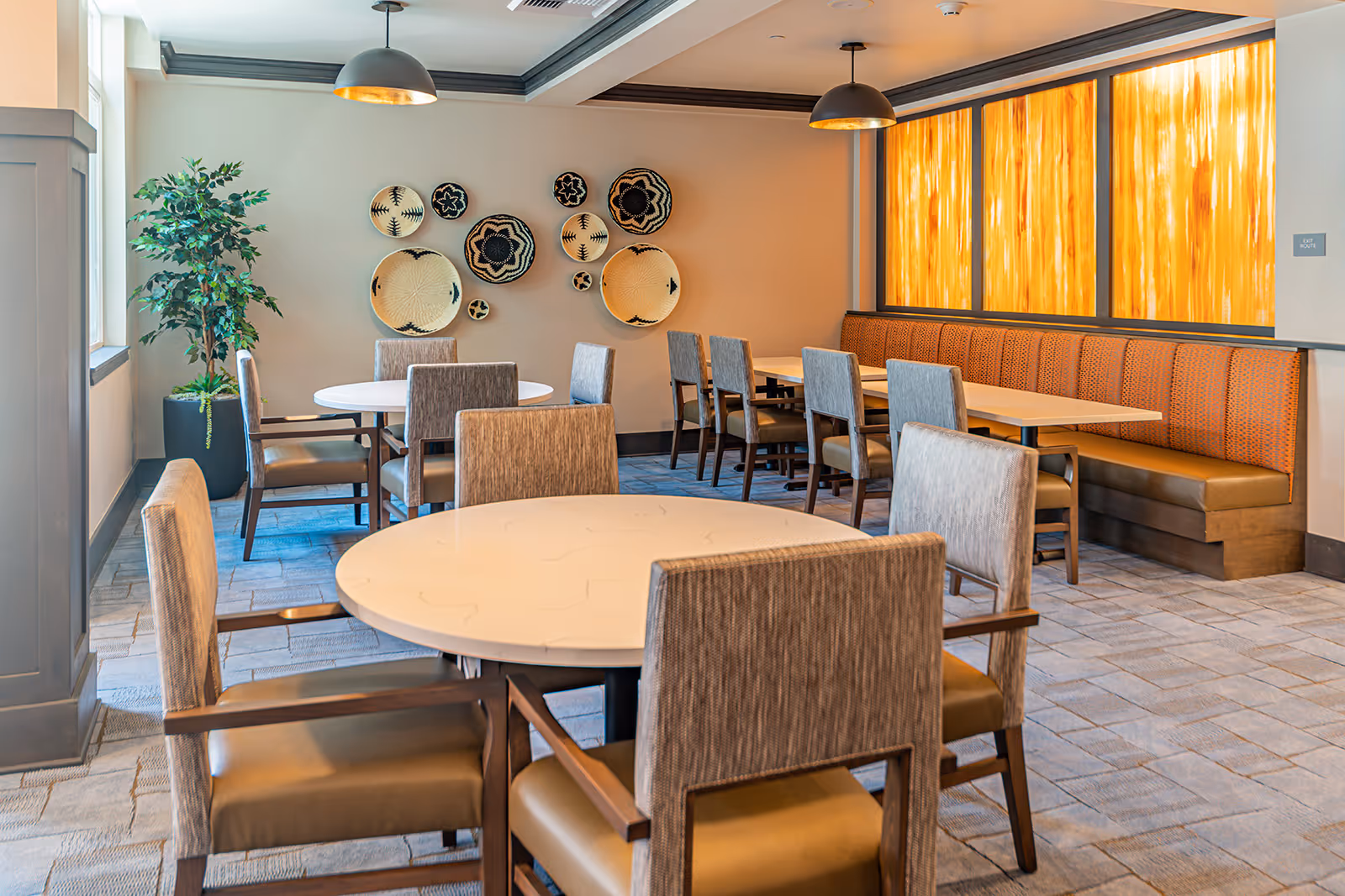 A bright dining room in a senior living facility with round and rectangular tables surrounded by upholstered chairs. The room features a decorative wall with woven baskets, a large potted plant near the window, and a long cushioned bench along one wall with warm amber backlighting.