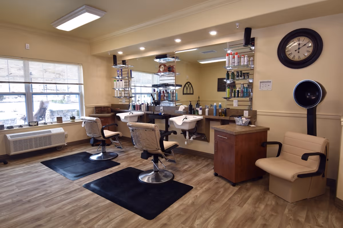 Interior of a salon area in a senior living facility with two salon chairs in front of sinks and mirrors, shelves stocked with hair care products, a beige salon dryer chair, a large wall clock, and a window with blinds letting in natural light.