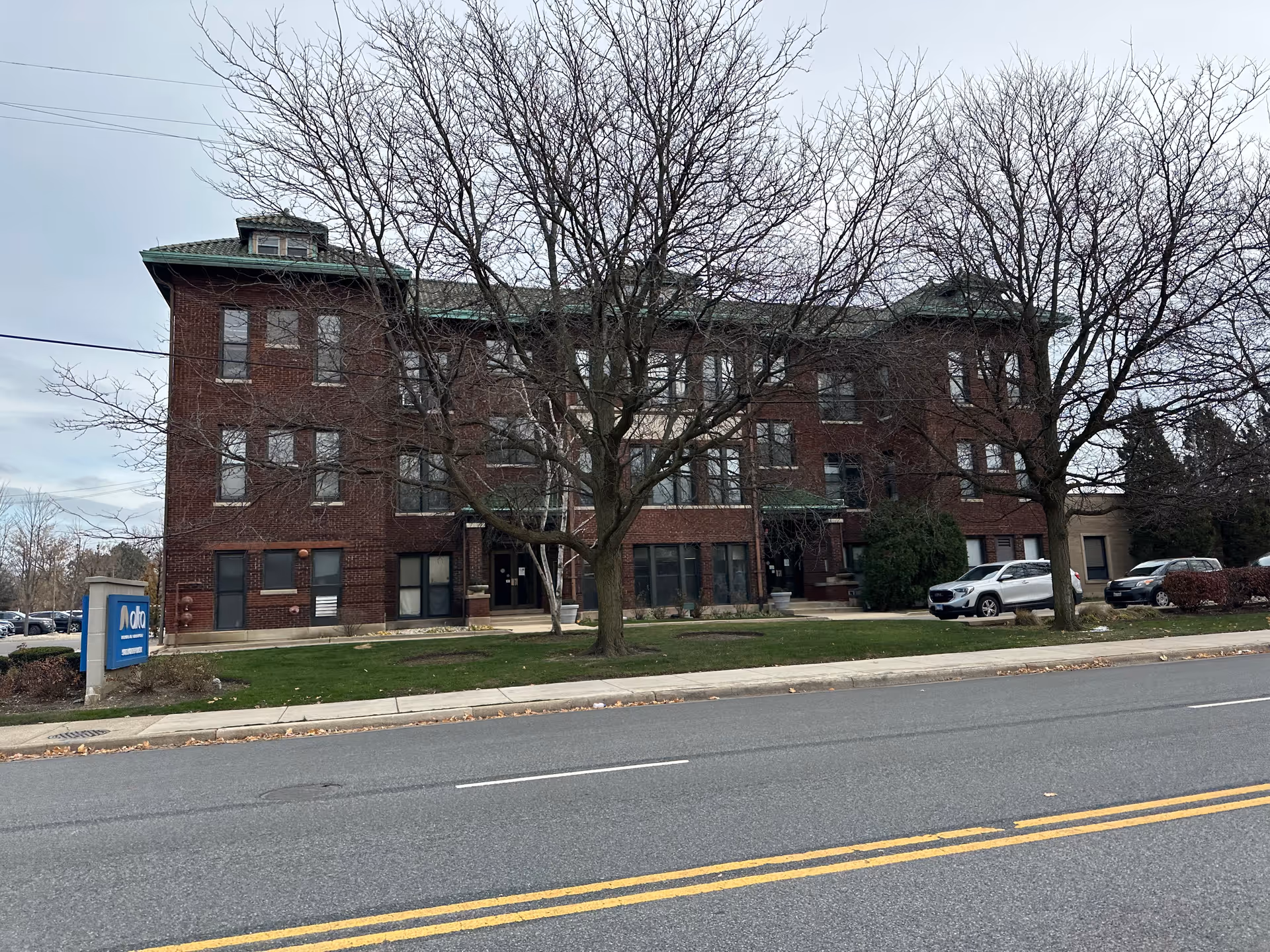 Exterior view of a three-story brick building with multiple windows and a green roof, situated behind leafless trees and a grassy area. There is a blue sign on the left side of the image that reads 'Alta Rehab at Fairmont.' Several cars are parked on the right side near the building, and a road with double yellow lines is visible in the foreground.