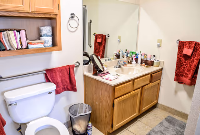 Bathroom with a toilet, wooden vanity and sink beneath a large mirror, red towels, and shelving with folded linens.