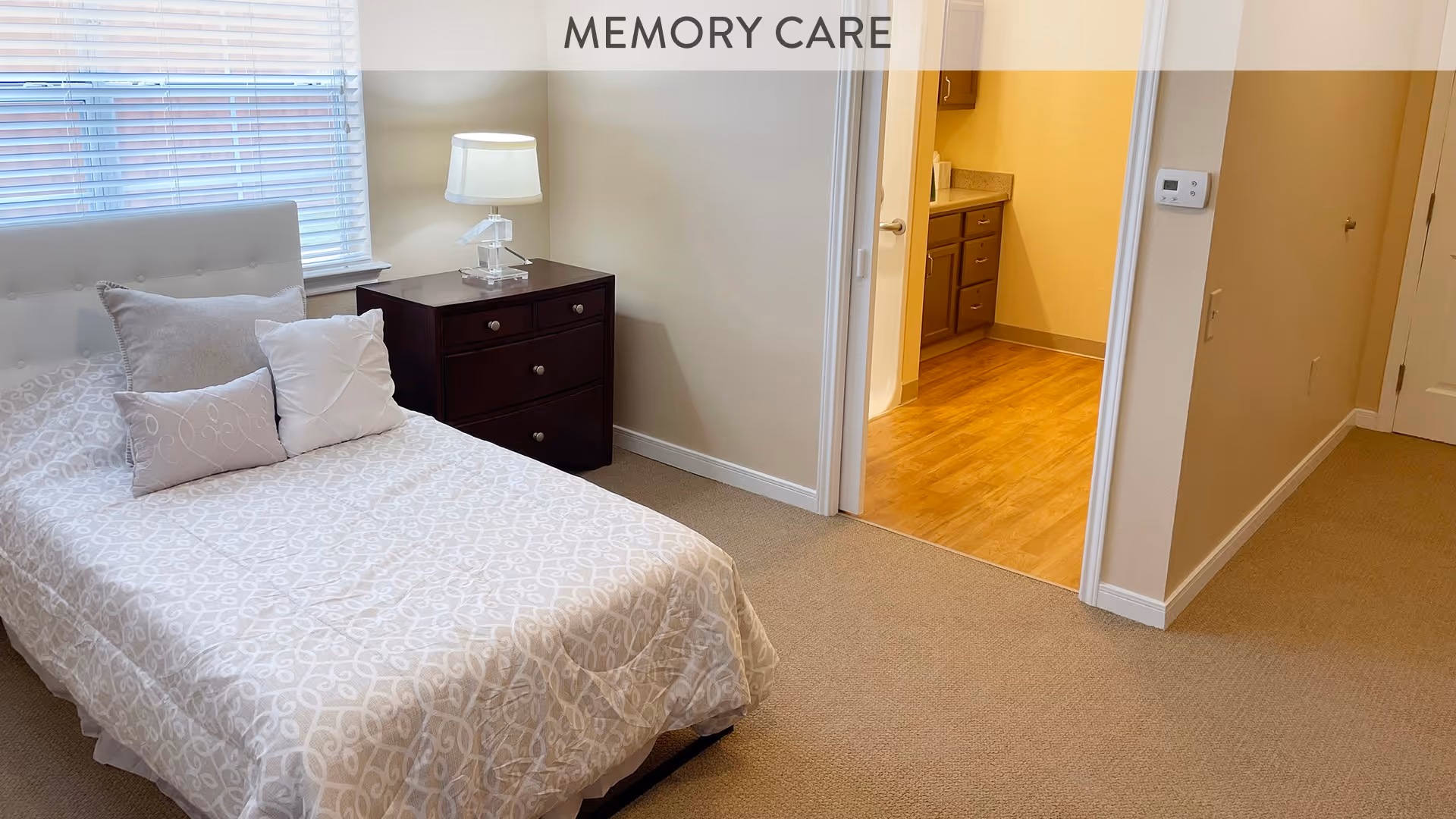 A cozy bedroom with a single bed covered in a light patterned bedspread and three pillows. Next to the bed is a dark wooden nightstand with a clear lamp on it. A window with blinds is on the left wall, and an open doorway leads to a room with wooden flooring and cabinetry. The word MEMORY CARE is displayed at the top center of the image.