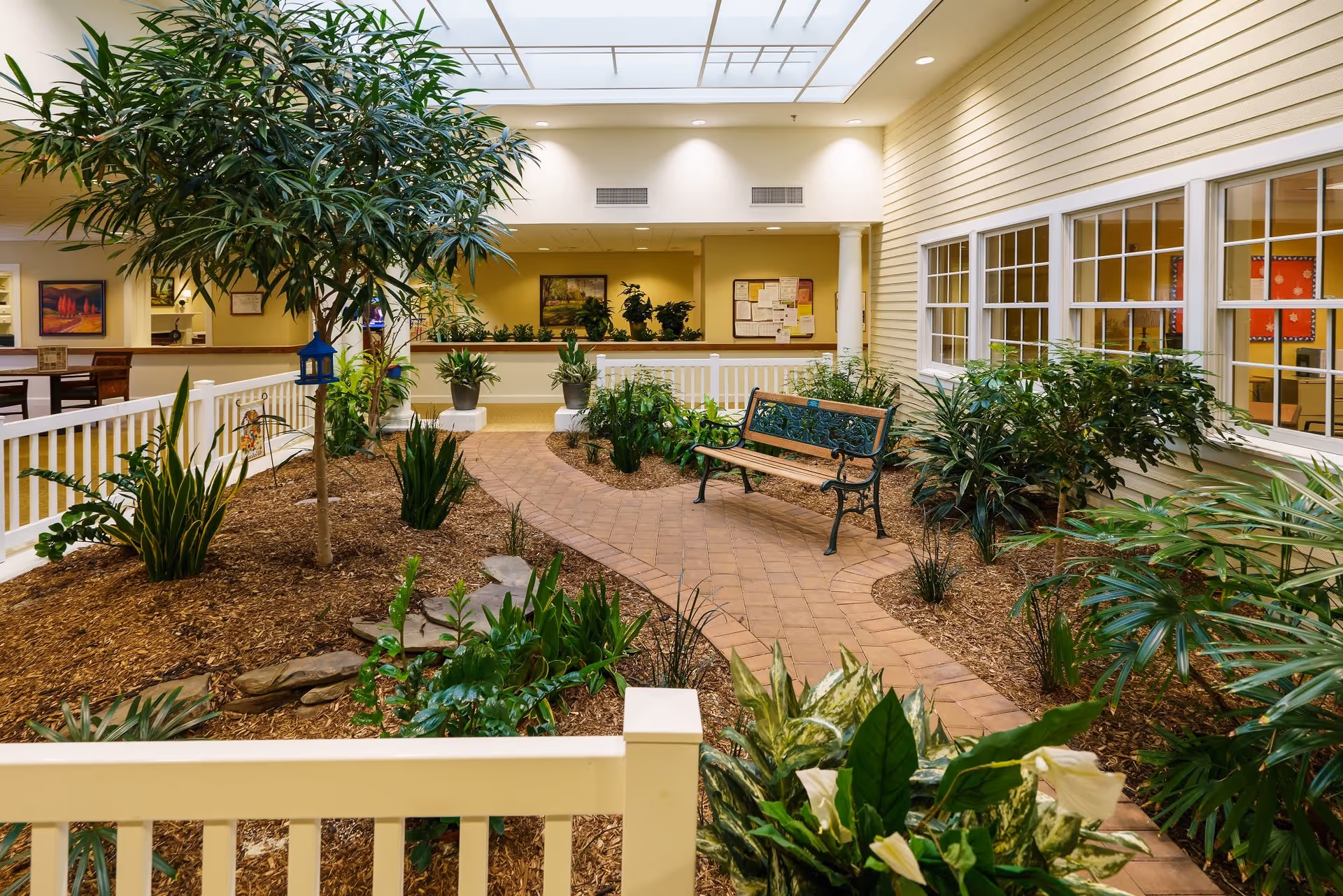 Indoor garden area in a senior living community with a brick pathway, green plants, a tree, and a decorative bench. The space is enclosed with white railings and has a skylight ceiling allowing natural light. There are windows and a bulletin board visible in the background.