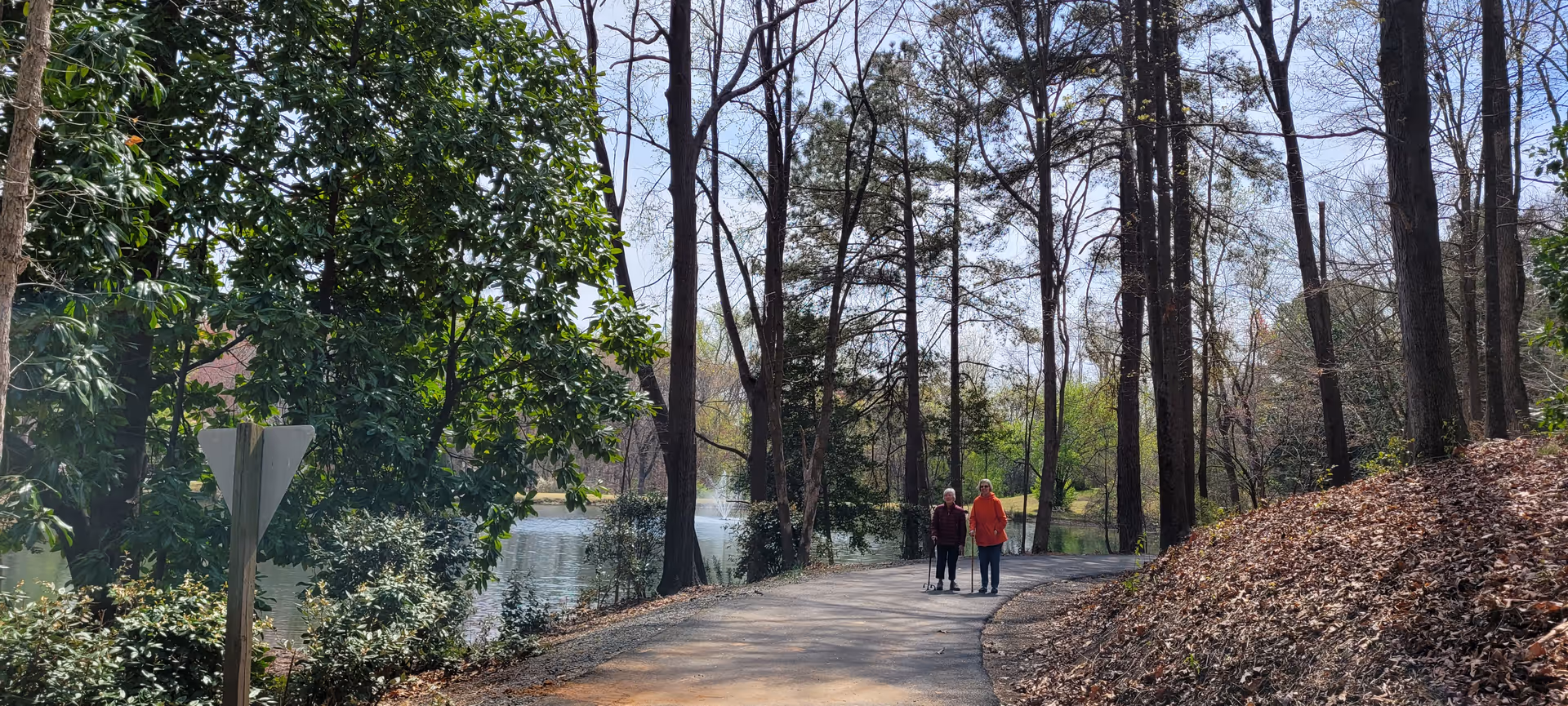 Two elderly people walking on a paved path surrounded by tall trees and greenery near a body of water with a fountain in the background on a clear day.