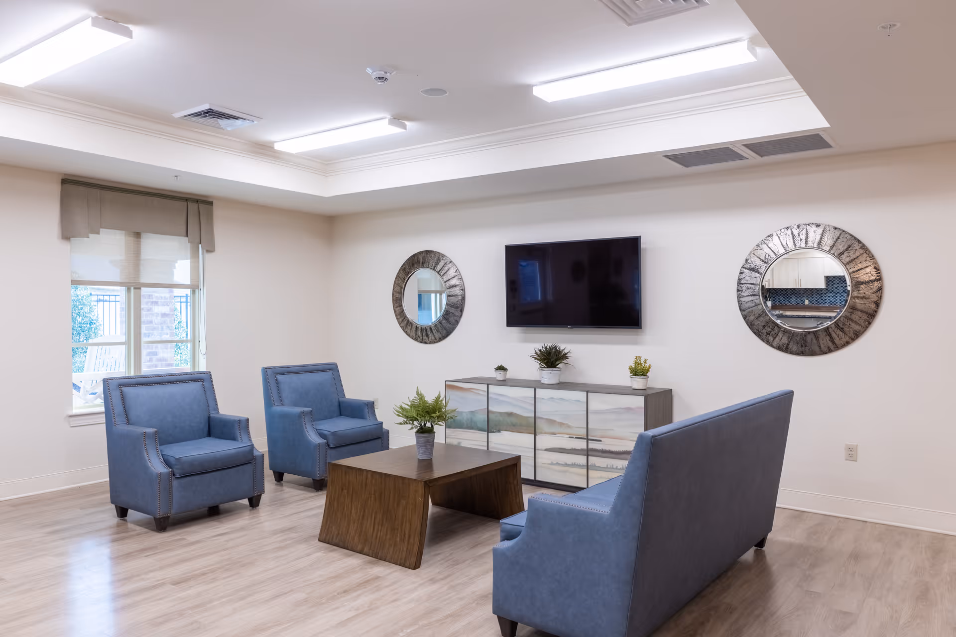 Bright communal living room with blue upholstered chairs and a sofa around a wooden coffee table facing a wall-mounted TV and decorative mirrors.