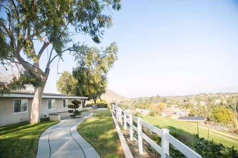 A curved sidewalk along a single-story building with a white fence and hillside view.