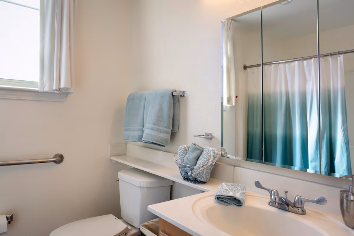 A clean and bright bathroom featuring a white toilet, a sink with a silver faucet, and a large mirror above the sink. Light blue towels hang on a towel rack above the toilet, and additional rolled towels are placed in a wire basket on the counter. A shower curtain with a gradient blue design is visible in the mirror reflection. A window with white curtains allows natural light into the room.