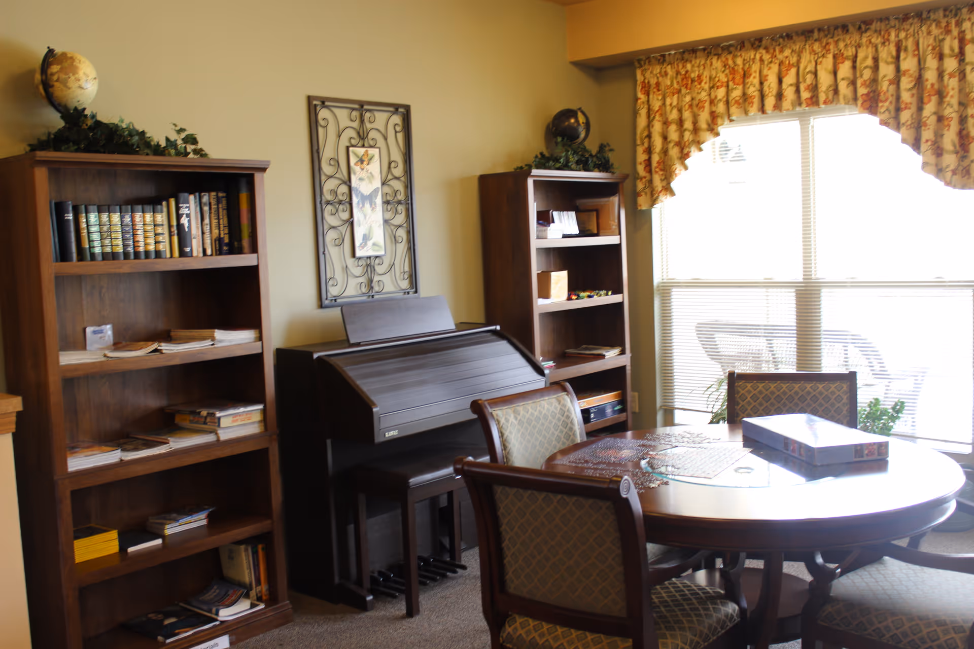 A cozy room with a round wooden table surrounded by four upholstered chairs. On the table, there is a partially completed jigsaw puzzle and a puzzle box. Behind the table, there are two wooden bookshelves filled with books and magazines, and a closed upright piano between them. A large window with floral curtains lets in natural light, and a patio with outdoor furniture is visible outside.