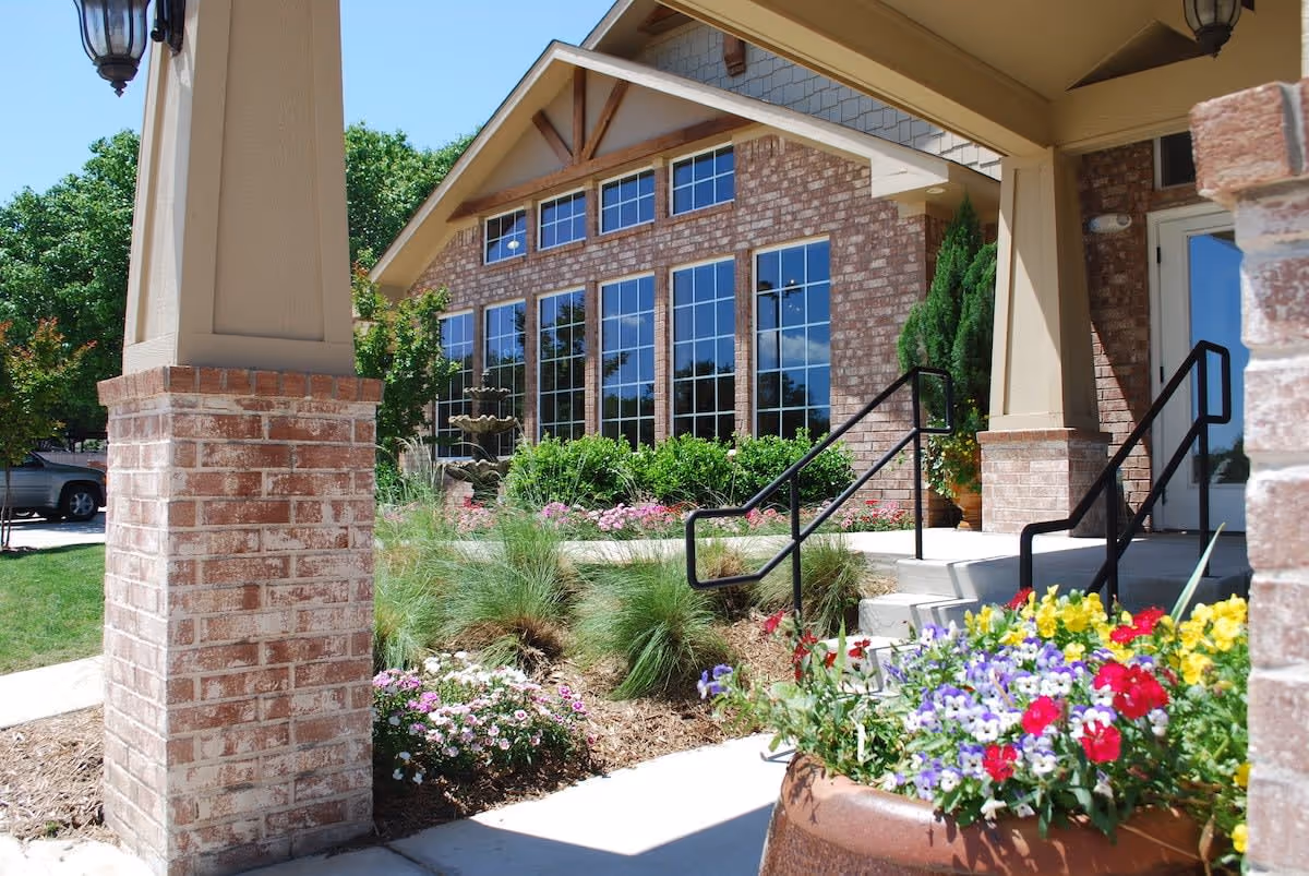 Exterior view of a brick building with large windows, surrounded by landscaped gardens with colorful flowers and greenery. There are steps with black handrails leading to a door, and a covered porch supported by brick columns.