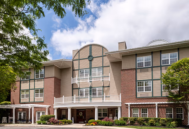 Exterior view of a three-story senior living facility building with a combination of brick and beige siding, white railings on balconies, and a covered entrance. The building is surrounded by green shrubs and trees under a partly cloudy sky.