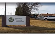 Outdoor view of a sign for Mennonite Friendship Communities with the mfc logo on a grassy area near a parking lot and some buildings in the background under a cloudy sky.