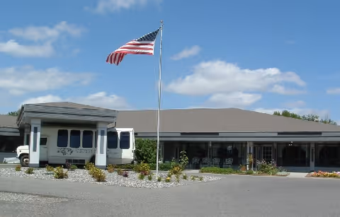 Front exterior view of Laurel Health and Rehabilitation Center building with a flagpole displaying the American flag in front, a covered entrance with a vehicle parked underneath, and a clear blue sky with some clouds.