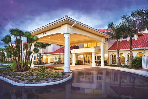 Exterior view of Discovery Village At The Forum showing the main entrance with a covered driveway, palm trees, and a building with red tile roofing under a cloudy sky at dusk.