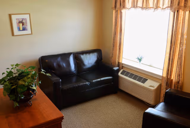 A small living room area with a black leather loveseat and a matching armchair. There is a wooden table with a green potted plant on it. A window with sheer curtains lets in natural light, and below the window is a wall-mounted air conditioning unit. A framed picture hangs on the beige wall.