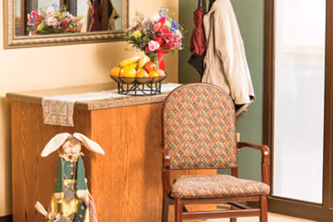 Entryway seating area with a patterned wooden armchair beside a wooden console topped with a fruit bowl and floral arrangement, coats hanging nearby.