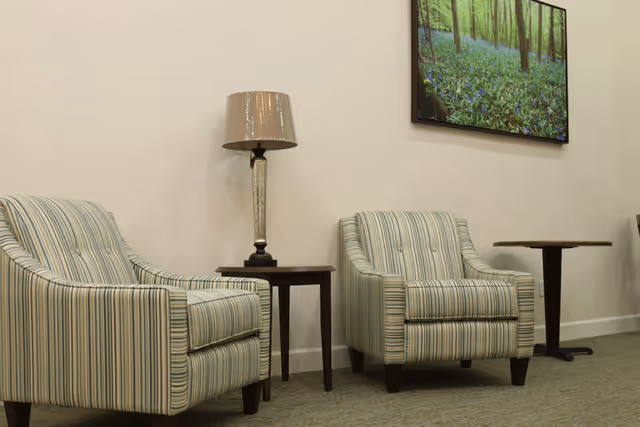 Two striped armchairs with a small wooden side table and a lamp between them, a small round table to the right, and a framed picture of a forest with green trees and blue flowers hanging on a beige wall.